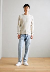 Beżowy sweter z warkoczy, z prążkowanymi mankietami, w połączeniu z jasnoniebieskimi spodniami prostymi i białymi sneakersami, stojący na drewnianej podłodze.