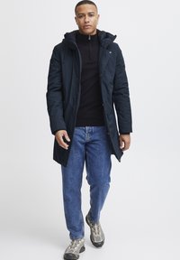 Marine isolert parka med hette, glidelåslukking og sidelommer. Kombinert med svart genser med høy hals, blå jeans og grå treningssko.