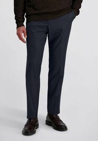 Man in een donkerblauwe op maat gemaakte broek, donkerbruine instappers en een donkerbruine trui, met één hand in zijn zak, staand tegen een effen achtergrond.