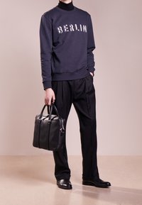 Felpa navy con testo "BERLIN", abbinata a pantaloni neri a righe sottili. Una borsa in pelle nera tenuta in mano, con doppi manici e hardware minimal.