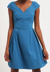 Eine Frau trägt ein figurbetontes, knielanges, blaues Kleid mit kurzen Ärmeln und einem V-Ausschnitt. Sie steht vor einem schlichten, hellen Hintergrund.