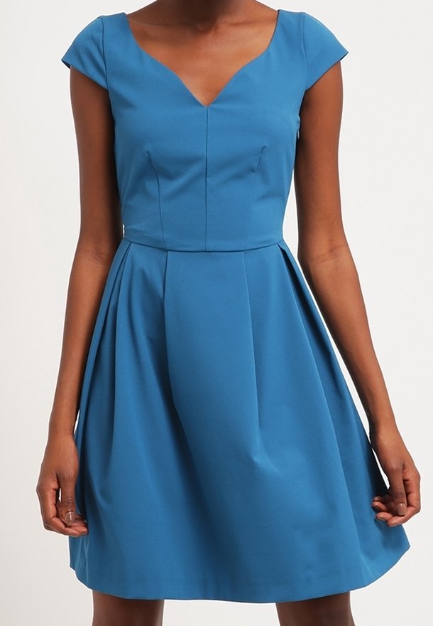 Eine Frau trägt ein figurbetontes, knielanges, blaues Kleid mit kurzen Ärmeln und einem V-Ausschnitt. Sie steht vor einem schlichten, hellen Hintergrund.