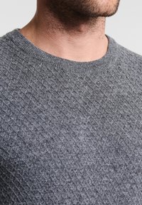 Grau gestrickter Pullover mit einem Rautenmuster, ausgestattet mit einem gerippten Ausschnitt und einer weichen Textur, gesehen aus dem oberen Brustbereich.