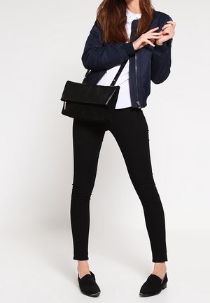 Veste bomber marine, t-shirt blanc, jean skinny noir, mocassins noirs et sac bandoulière noir, avec des fermetures éclair et un design minimaliste.