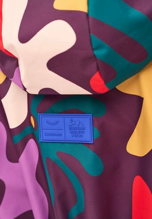 Kolorowa tkanina z abstrakcyjnymi kształtami w kolorach fioletowym, zielonym, żółtym, białym i czerwonym, z niebieską metką z napisem "Naoko Explorers" oraz tekstem dotyczącym dziczy.