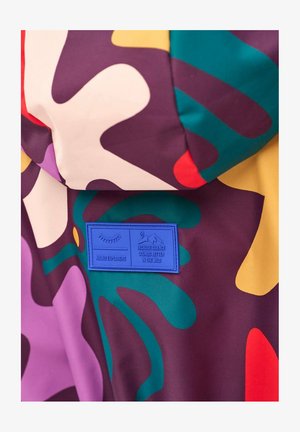 Kolorowa tkanina z abstrakcyjnymi kształtami w kolorach fioletowym, zielonym, żółtym, białym i czerwonym, z niebieską metką z napisem "Naoko Explorers" oraz tekstem dotyczącym dziczy.