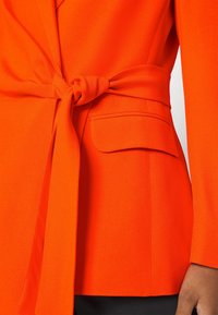 Blazer orange vif avec une ceinture nouée à la taille et une poche à rabat, porté par une personne avec une main visible et un pantalon foncé.