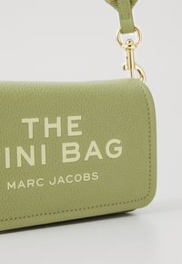 Roheline nahast mini kott kuldsete detailidega. Omab reljeefset teksti: "THE MINI BAG" ja "MARC JACOBS". Struktureeritud kuju ja tekstureeritud viimistlus.