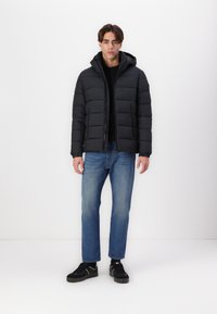 Zwarte pufferjack met capuchon, voorzien van een rits en een doorgestikt ontwerp. Gecombineerd met blauwe denim jeans en zwarte sneakers.