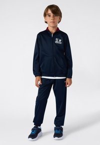 Granatowy dres wykonany z gładkiego materiału. Kurtka ma pełny zamek błyskawiczny i dwie przednie kieszenie. Połączony z dopasowanymi spodniami z elastycznymi mankietami.