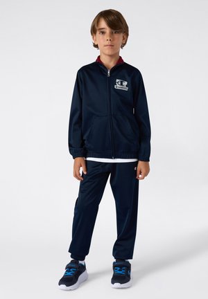 Granatowy dres wykonany z gładkiego materiału. Kurtka ma pełny zamek błyskawiczny i dwie przednie kieszenie. Połączony z dopasowanymi spodniami z elastycznymi mankietami.