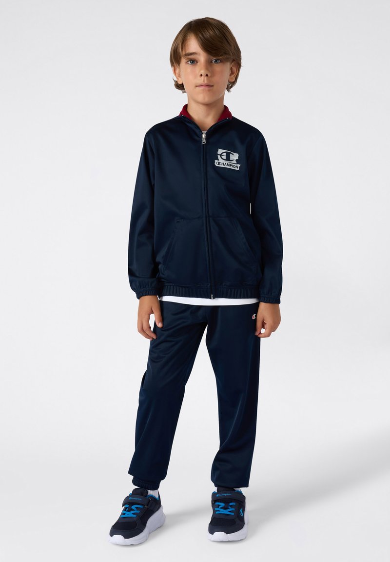 Granatowy dres wykonany z gładkiego materiału. Kurtka ma pełny zamek błyskawiczny i dwie przednie kieszenie. Połączony z dopasowanymi spodniami z elastycznymi mankietami.