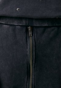 Sort lynlåsgarment med en glat tekstur, der har en fremtrædende metallynlås og trykknapper øverst, der viser et minimalistisk design.