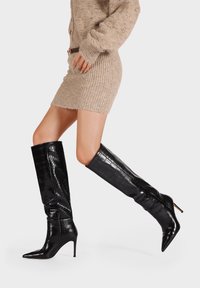 Stivali neri al ginocchio in pelle di coccodrillo, con punta affilata e tacco a spillo, abbinati a un vestito in maglia beige.
