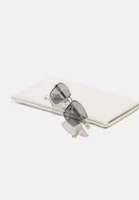 Lunettes de soleil avec des verres gris et une monture argentée, posées en angle sur une pochette en cuir souple blanc avec des coutures discrètes et une finition lisse.