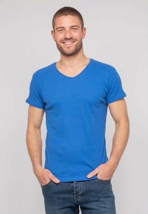 T-shirt bleu à col en V fabriqué en tissu doux, avec des manches courtes et une coupe ajustée. Porté avec un jean bleu décontracté, les mains dans les poches.