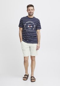 Marinblå stripete t-skjorte med grafisk design, lyse beige shorts og brune sandaler. Uformell antrekk med korte ermer og løs passform.