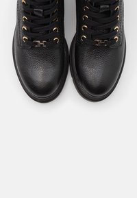 Mustat tekstuuripintaiset nahkasaappaat, joissa on kultaiset nauhakengät ja metalliset alut mustissa nauhoissa, nähtynä ylhäältä päin valkoista taustaa vasten.