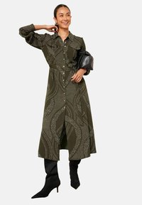 Rochie cămașă verde măsliniu cu model de valuri albe, cu nasturi în față, buzunare și talie ajustată. Asortată cu cizme negre până la genunchi cu vârf ascuțit.