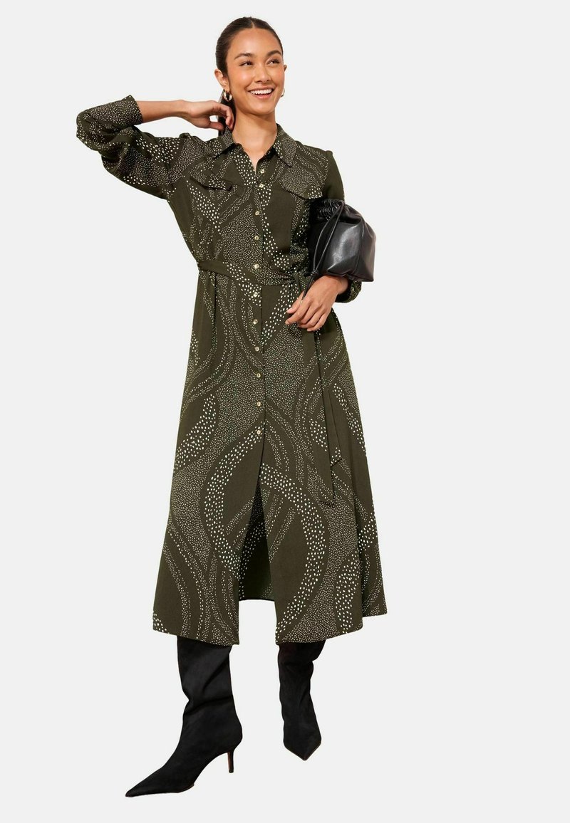 Rochie cămașă verde măsliniu cu model de valuri albe, cu nasturi în față, buzunare și talie ajustată. Asortată cu cizme negre până la genunchi cu vârf ascuțit.