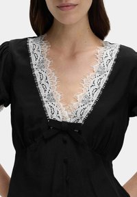 Schwarzes Kleid mit tiefem V-Ausschnitt, weißer Spitze, Schleifendetail in der Taille und Knopfverschluss auf der Vorderseite.