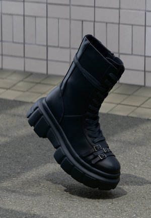 Schwarzer klobiger Kampfstiefel mit dicker Sohle, Schnürung vorne, doppelte Schnallenriemen nahe dem Zehenbereich, gezeigt vor einem gefliesten Wand- und Gehweghintergrund.
