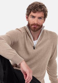 Beiger Strickpullover mit Reißverschluss und Stehkragen, gerippten Bündchen und weicher Textur. Wird über einem weißen Hemd getragen und mit dunkler Hose kombiniert.