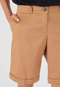 Pantalones cortos caqui hechos de una tela suave y tejida con un corte recto. Cuentan con bolsillos laterales y un dobladillo enrollado, asegurado con un cierre de botón.