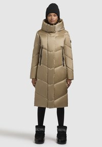 Beige quilted puffer coat with a high collar and zippered side pockets. Worn with black leggings and black fur-lined boots. Solid color texture.