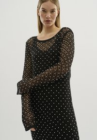 Rochie tricotată neagră cu un model din plasă, decorată cu perle albe, cu mâneci lungi și decolteu în formă de inimă.