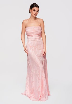 Donna con capelli scuri in un elegante abito lungo fino a terra color rosa chiaro con spalline sottili e motivi floreali delicati su sfondo bianco.