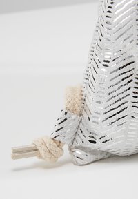 Gros plan d’un coin de sac argenté à motif avec une corde tressée crème nouée, terminée par deux embouts argentés.