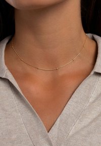 Goldene Halskette mit einem filigranen Design, kleinen runden Akzenten, feiner Textur und schlankem Profil, die am Hals getragen wird.