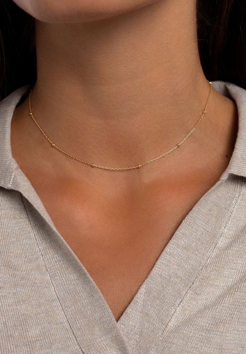 Goldene Halskette mit einem filigranen Design, kleinen runden Akzenten, feiner Textur und schlankem Profil, die am Hals getragen wird.