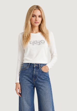 Mujer rubia con una camiseta blanca de manga larga con un logo y vaqueros azules de talle alto y pata ancha, posando con una mano en el bolsillo.