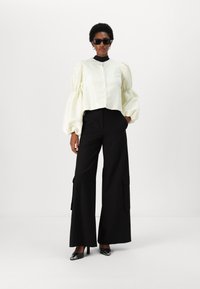 Blouse courte en satin ivoire avec des manches bouffantes, associée à un pantalon noir taille haute à jambes larges avec poches cargo, le tout terminé par des talons noirs.