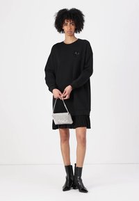 Sweatshirt noir surdimensionné à manches longues, avec un petit logo rehaussé. Porté par-dessus une jupe plissée noire et accompagné de bottines.