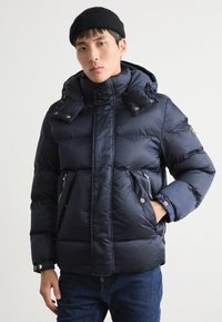 Marinblå pufferjacka med texturerad yta, hög krage, avtagbar huva, dubbla dragkedjefickor och knappknäppning på axlarna.