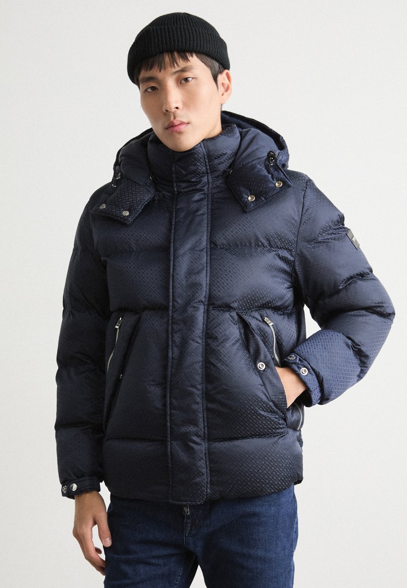 Marinblå pufferjacka med texturerad yta, hög krage, avtagbar huva, dubbla dragkedjefickor och knappknäppning på axlarna.