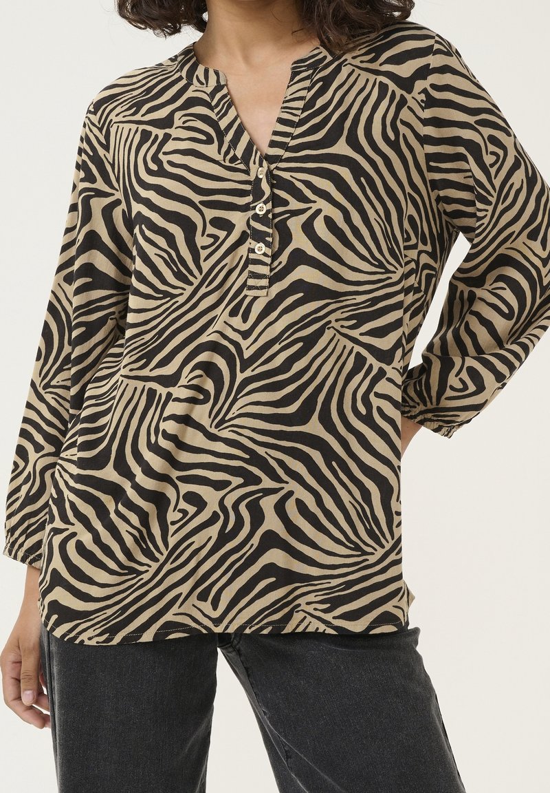 Blouse beige avec imprimé zèbre noir, dotée d'un col en V, d'une patte à quatre boutons et de manches trois-quarts amples. Texture de tissu douce.