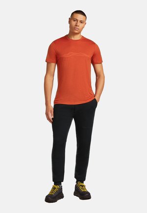 Man staat in een roestoranje T-shirt met korte mouwen, zwarte broek en zwarte wandelschoenen met gele veters voor een witte achtergrond.