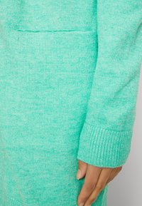 Détail d'un pull en tricot vert menthe montrant un tissu texturé, une manchette côtelée et une poche kangourou à l'avant. Doux, de medium épaisseur.