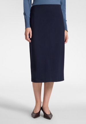 Mujer con falda azul marino hasta la rodilla, top azul de manga larga, zapatos oscuros puntiagudos, de pie con las manos relajadas a los lados.