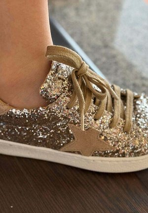 Pie de niño con zapatilla brillante dorada con cordones beige y aplique de estrella, parado sobre un suelo de madera oscuro.