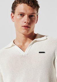 Tricou polo alb tricotat, cu guler în V și un model texturat din plasă. Prezintă un accent cu logo negru pe piept și un guler de contrast.