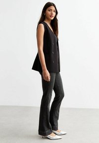 Blazer sans manches noir avec un décolleté en V et des boutons double boutonnage, associé à un jean flare noir et des escarpins blancs à bout pointu.