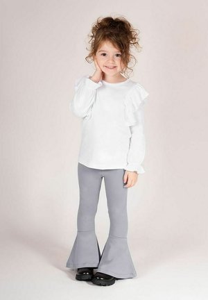 Biała bluzka z długim rękawem ozdobiona falbankami oraz szare legginsy dzwony. Dziecko w czarnych butach stoi, opierając rękę na policzku.