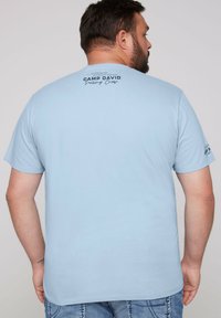 Camiseta de manga corta azul claro con dobladillo recto y un logo impreso en la parte posterior. El material parece suave y similar al algodón.