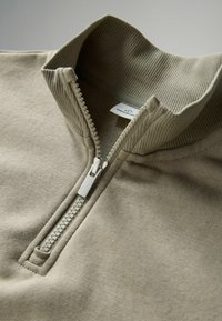 Half-zip pullover kevyessä beige-puuvillasekoitteessa. Röyhelö-kaulus ja pehmeä, teksturoitu kangas, jossa on hopeinen vetoketju ja ompeluyksityiskohtia.