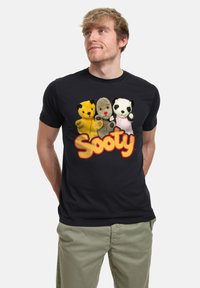 Černé bavlněné tričko s krátkým rukávem a kulatým výstřihem, na kterém jsou čtyři plyšoví loutkoví hrdinové a text "Sooty" tučně napsaný žlutooranžovým písmem.